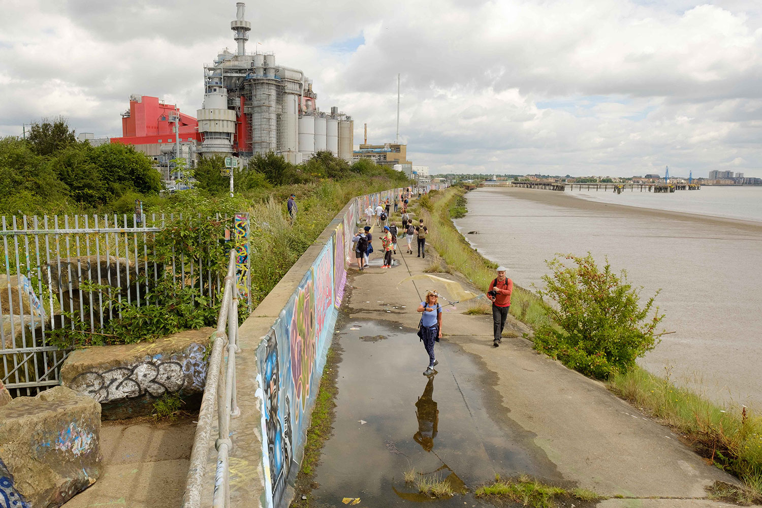 Walkers approach along the graffiti wall between a large factory on the left and the Thames river on the right